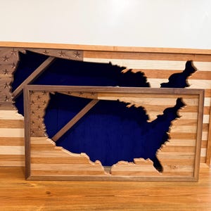 May include: Two wooden wall art pieces depicting the United States map, with a flag design. The larger piece is rectangular, and the smaller one is square. The background of the map is blue, and the frame is made of wood.