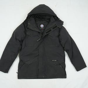 May include: Black hooded winter jacket with multiple pockets and a zipper closure. The jacket features a logo patch on the chest and a hood. The jacket is designed for cold weather and outdoor activities.