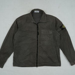 May include: A dark gray long-sleeve shirt with a collared neckline and a full-length black zipper. The shirt has a single chest pocket and a small patch with a green logo on the left sleeve.