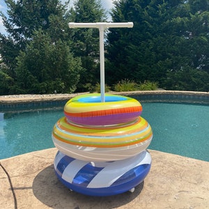 May include: A white PVC pipe stand with a T-shaped top holds three colorful inflatable pool rings. The rings are stacked on top of each other, with the largest ring at the bottom and the smallest ring at the top. The rings are blue, yellow, orange, and purple.