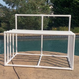 May include: White PVC pipe frame structure with multiple horizontal bars. The frame is sitting on a concrete patio near a swimming pool.