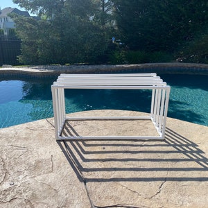 May include: White metal frame with a rectangular shape, sitting on a stone surface near a swimming pool.