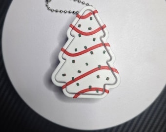 May include: A white Christmas tree-shaped keychain with a silver ball chain. The tree features raised red diagonal stripes and small green rectangular accents scattered across the surface, resembling a decorated holiday cookie.