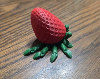 May include: A red, textured strawberry-shaped object attached to a green, multi-legged base that resembles a spider or crab. The strawberry has a pitted surface, and the green base features several articulated, segmented appendages.