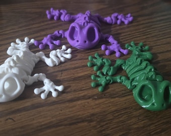 May include: Three articulated frog figurines in white, purple, and green. Each frog features a skeletal-like structure with large eye sockets, segmented limbs, and detailed toes, designed with a flexible, jointed body construction.