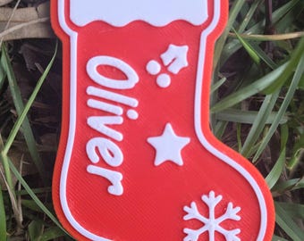 May include: A red and white Christmas stocking ornament featuring the name Oliver in raised white lettering. The stocking is decorated with a white holly sprig, a five-pointed star, and a snowflake icon, with a hole at the top for hanging.