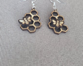 May include: A pair of dangle earrings featuring laser-cut wooden honeycomb shapes with a small bee silhouette attached to the front. The earrings are suspended from silver-toned metal hooks, showcasing a natural brown wood finish.