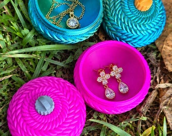 May include: Two round, textured trinket boxes with lids. One box is teal with an orange knob and contains a gold chain necklace with a round pendant. The other box is bright pink with a gray knob and contains two gold flower-shaped earrings.