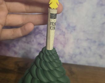 May include: A dark green, cone-shaped object with a spiraling, layered texture resembling a stylized tree. A rolled-up paper bill is inserted into the top center, topped with a small, bright yellow, five-pointed star ornament with a hole.