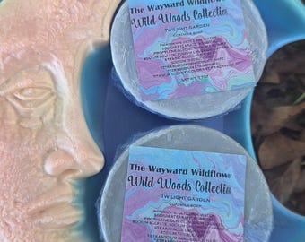 May include: Two round bars of Twilight Garden goat milk soap from The Wayward Wildflower Wild Woods Collection, each wrapped in plastic with a label, resting inside a crescent moon-shaped dish with a peach and light blue gradient finish.