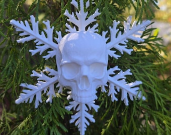 May include: A white, three-dimensional ornament featuring a central human skull integrated into a six-pointed snowflake design. The object has a smooth, matte finish and includes a small circular loop at the top for hanging purposes.