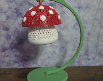 May include: A 3D printed mushroom-shaped object with a red lattice cap featuring white dots and a white lattice stem. The mushroom hangs from a curved green stand attached to a circular green base, creating a hanging display structure.