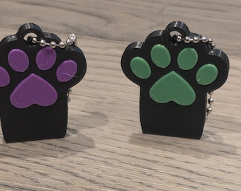 May include: Two black paw-shaped keychains with metal ball chains. One keychain features purple paw pads, while the other features green paw pads. Both items have a hole at the top for attachment and a textured surface on the colored paw pads.