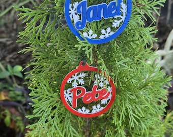 May include: Two circular ornaments featuring white snowflake cutouts. The top ornament is blue with the name Janet in blue script. The bottom ornament is red with the name Pete in red script. Both ornaments hang from green hooks on a green tree.