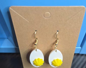 May include: A pair of dangle earrings featuring a white oval base with a textured yellow center, resembling a fried egg. The earrings are attached to gold-toned metal hooks and are displayed on a tan cardboard jewelry display card.