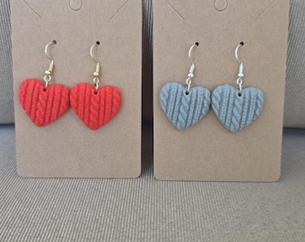 May include: Two pairs of heart-shaped dangle earrings displayed on brown cardboard cards. One pair is bright red with a textured cable knit pattern and gold-toned hooks, while the other pair is light gray with the same texture and silver-toned hooks.