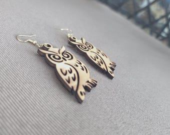 May include: A pair of dangling earrings featuring laser-cut wooden owl shapes. The owls are light tan with dark brown engraved details for the eyes, feathers, and wings. Each earring is attached to a simple silver-toned metal fishhook wire.