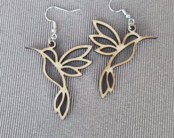 May include: A pair of dangle earrings featuring laser-cut wooden hummingbird silhouettes. Each earring has a light brown wood grain finish with dark edges and is attached to a silver-toned metal fishhook ear wire for hanging.