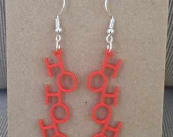 May include: A pair of dangling earrings featuring the red text HO HO HO stacked vertically. The letters are constructed from a textured material and hang from silver-toned metal fishhook wires attached to a brown cardboard display card.