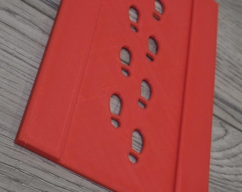 May include: A rectangular red plastic stencil featuring a series of small, cut-out footprints arranged in a vertical path down the center. The stencil has raised edges on both the left and right sides, providing a structured frame for the design.