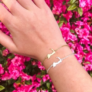 May include: A gold and silver bracelet with a charm shaped like the state of Massachusetts. The bracelet is worn on a person's wrist against a background of pink flowers.