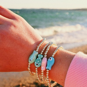 May include: Four beaded bracelets with colorful fish charms. The fish charms are light blue, mint green with white polka dots, pink, and blue. The bracelets are gold-colored and are worn on a wrist near the ocean.