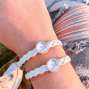 May include: Two white braided bracelets with a round charm featuring a map of an island. The charms are silver with a white background and a blue outline of the island.