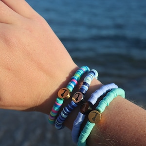 May include: A set of four colorful beaded bracelets with gold charms. The bracelets are stacked on a person's wrist. The charms are shaped like a small island and have the letter "T" engraved on them.