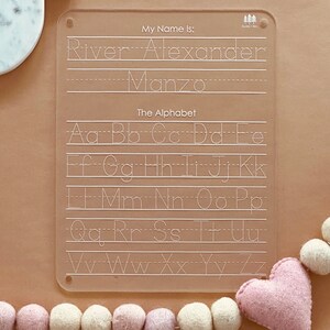 May include: A clear plastic dry-erase board with a dotted alphabet and lines for writing. The board has the text "My Name Is:" and "The Alphabet" printed on it. The board is on a brown surface with a pink heart and white and pink balls.