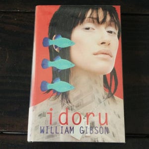 May include: A book cover featuring a woman's face with three turquoise fish illustrations. The title "idoru" is in red, with the author's name, William Gibson, below. The background is a mix of red and a faded image.