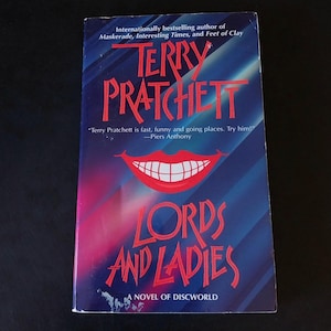 May include: A paperback book titled Lords and Ladies by Terry Pratchett, a novel of Discworld. The cover features a dark blue and purple gradient background with red text and a graphic of a wide, smiling mouth with white teeth in the center.