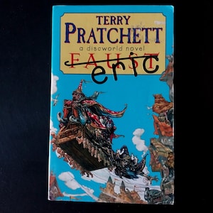 May include: A paperback book titled "Eric" by Terry Pratchett, a discworld novel. The cover features a vibrant illustration of a wizard on a flying platform against a blue sky background. The title "Faust" is crossed out.