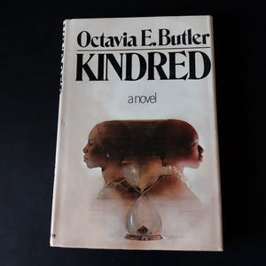 May include: A paperback book titled Kindred by Octavia E. Butler. The white cover features black text and an illustration of two profiles facing away from each other, separated by a central hourglass graphic with a soft, faded brown background.