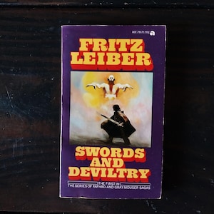 Op de afbeelding: Een paars paperback boek met een gele en rode titel, "Fritz Leiber Swords and Deviltry", de eerste in de serie van Fafhrd en Gray Mouser Sagas. De omslag toont een fantasievolle illustratie van een man met een zwaard die voor een gloeiende figuur staat.