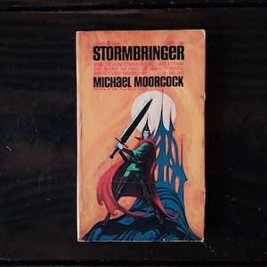 May include: A paperback book cover with a yellow, orange, and blue illustration of a man in a green tunic and red cape holding a sword. The book title is "Stormbringer" and the author is "Michael Moorcock".