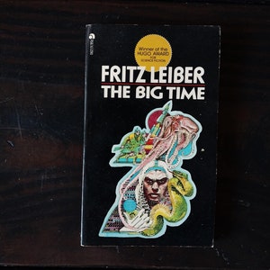 Op de afbeelding: Een zwart paperback boek met een kleurrijke illustratie van een wezen met tentakels en een menselijk gezicht. De titel van het boek is "Fritz Leiber The Big Time" en de omslag vermeldt "Winner of the Hugo Award for Science Fiction".