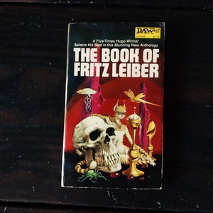 Op de afbeelding: Een paperback boekomslag met een rode, witte en zwarte illustratie van een schedel, een katachtig wezen en een gouden kelk. De titel van het boek is "The Book of Fritz Leiber".