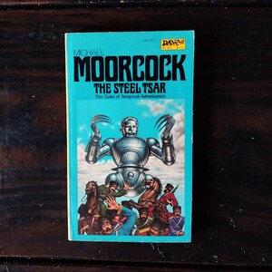 Könnte beinhalten: Ein blaues Taschenbuch mit dem Titel "Der Stahlzar" von Michael Moorcock. Das Cover zeigt einen silbernen Roboter mit einem bedrohlichen Ausdruck und eine Gruppe von Menschen auf Pferden.