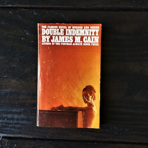 Può includere: Un libro tascabile vintage intitolato "Double Indemnity" di James M. Cain. La copertina presenta una donna e uno sfondo sfumato arancione e giallo. Il testo recita "Il famoso romanzo di omicidio e desiderio."