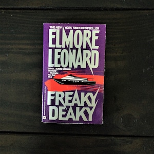 Puede incluir: Un libro de bolsillo titulado "Freaky Deaky" de Elmore Leonard. La portada es morada con texto blanco y una ilustración roja y negra de un barco. El texto dice "The New York Times Bestseller".