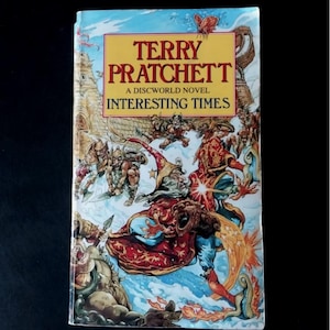 May include: A paperback book titled "Interesting Times" by Terry Pratchett. The cover features colorful fantasy illustrations of wizards, warriors, and other characters. The book is a Discworld novel.