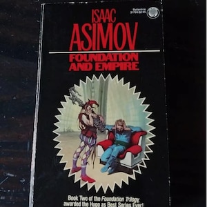Puede incluir: Un libro de bolsillo negro con un título rojo y blanco, "Isaac Asimov Fundación e Imperio". La portada presenta una ilustración de dos hombres en un entorno futurista. El libro forma parte de la Trilogía de la Fundación y fue galardonado con el premio Hugo a la mejor serie de todos los tiempos.