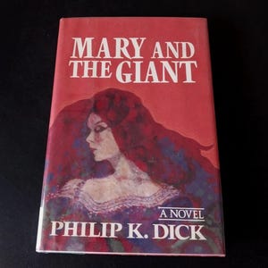 以下が含まれることがあります： フィリップ・K・ディック著の「MARY AND THE GIANT」というタイトルのヴィンテージペーパーバック。表紙は赤を基調とし、長い赤毛の女性の肖像画が描かれています。タイトルは白で、著者の名前は下部にあります。