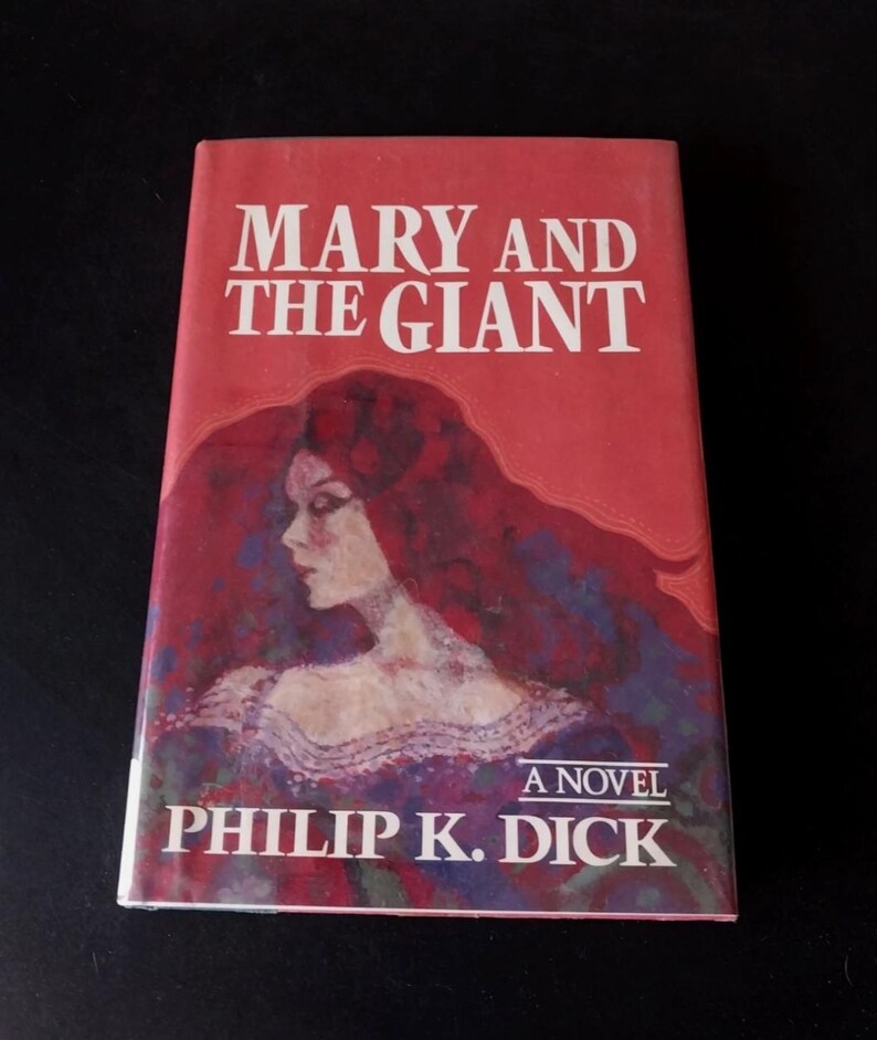 以下が含まれることがあります： フィリップ・K・ディック著の「MARY AND THE GIANT」というタイトルのヴィンテージペーパーバック。表紙は赤を基調とし、長い赤毛の女性の肖像画が描かれています。タイトルは白で、著者の名前は下部にあります。