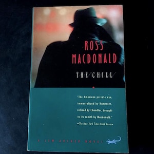 Può includere: Un libro tascabile intitolato "The Chill" di Ross Macdonald. La copertina presenta una figura in ombra con un cappello su uno sfondo sfocato. Il titolo e il nome dell'autore sono in rosso e bianco. È visibile anche una citazione dal New York Times Book Review.