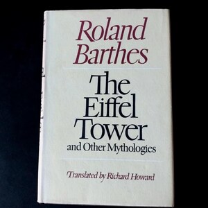 May include: A book with a cream-colored cover featuring the title The Eiffel Tower and Other Mythologies by Roland Barthes in black and maroon serif text. The text Translated by Richard Howard appears in smaller black italics at the bottom.