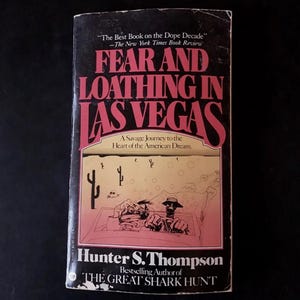 May include: A paperback book titled Fear and Loathing in Las Vegas by Hunter S. Thompson. The black cover features pink text and a line drawing of two figures in a convertible driving through a desert landscape with cacti and circular shapes.