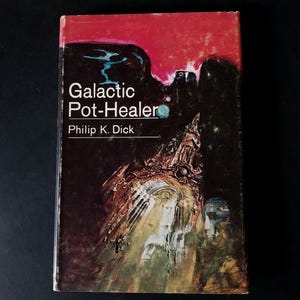 Może przedstawiać: Książka w miękkiej oprawie w stylu vintage zatytułowana "Galactic Pot-Healer" autorstwa Philipa K. Dicka. Okładka przedstawia czerwono-czarny abstrakcyjny wzór z futurystyczną ilustracją w odcieniach brązu, zieleni i błękitu. Książka jest na czarnym tle.