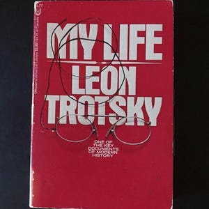 May include: A red book titled My Life by Leon Trotsky with white text and the subtitle One of the Key Documents of Modern History. A pair of thin, silver-rimmed pince-nez glasses with a black cord rests on the front cover of the book.