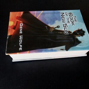 Puede incluir: Un libro de tapa dura titulado "The Book of the New Sun" de Gene Wolfe. La portada presenta una figura con una capa oscura y una espada, sobre un cielo azul y naranja. El libro est&aacute; sobre una superficie negra.